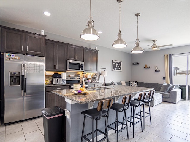 kitchen featuring stainless steel appliances, dark brown cabinetry, a center island with sink, decorative light fixtures, and dark stone counters