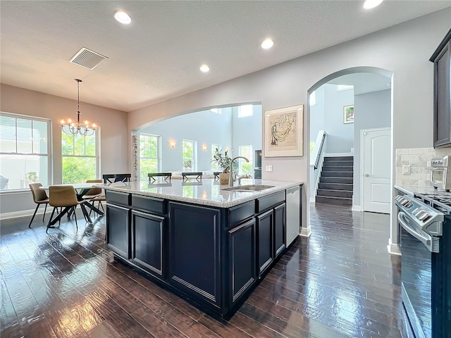 kitchen with sink, a kitchen island with sink, hanging light fixtures, stainless steel appliances, and light stone counters