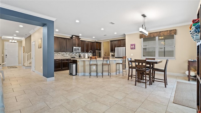 kitchen with hanging light fixtures, backsplash, dark brown cabinets, stainless steel appliances, and a center island with sink