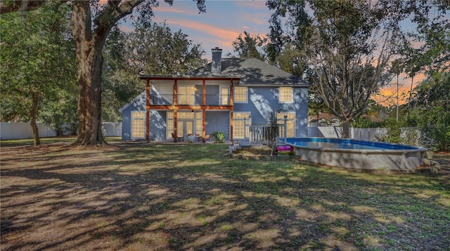 back house at dusk featuring a fenced in pool, a balcony, french doors, and a lawn