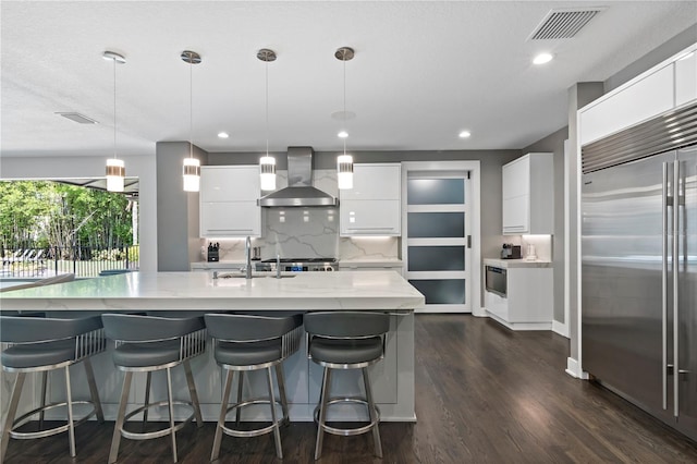 kitchen featuring white cabinetry, hanging light fixtures, an island with sink, stainless steel built in fridge, and wall chimney exhaust hood