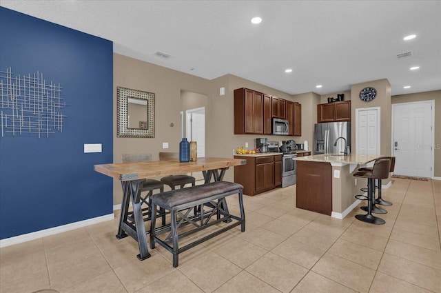 kitchen featuring appliances with stainless steel finishes, a kitchen breakfast bar, light tile patterned floors, light stone counters, and a center island with sink