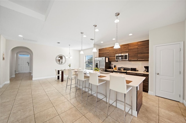 kitchen featuring tasteful backsplash, hanging light fixtures, a center island with sink, light tile patterned floors, and stainless steel appliances