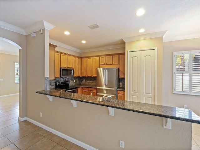 kitchen featuring light tile patterned flooring, appliances with stainless steel finishes, dark stone countertops, a breakfast bar area, and kitchen peninsula