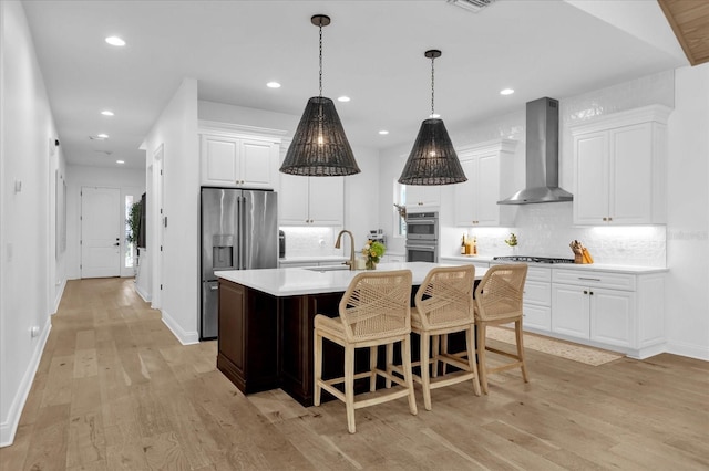 kitchen featuring wall chimney range hood, appliances with stainless steel finishes, white cabinetry, an island with sink, and light wood-type flooring