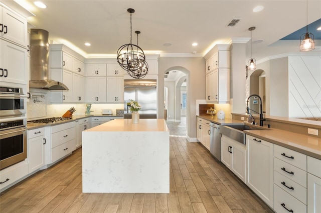 kitchen with sink, decorative light fixtures, wall chimney range hood, stainless steel appliances, and white cabinets