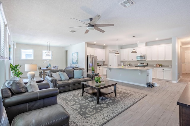 living room featuring light wood-style flooring, ceiling fan with notable chandelier, visible vents, and a textured ceiling