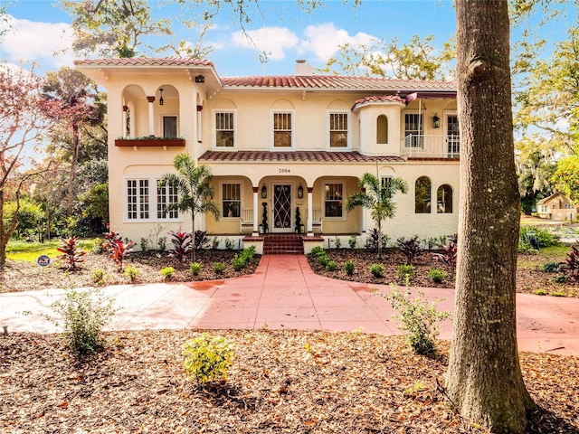 mediterranean / spanish house with a balcony, covered porch, a tile roof, stucco siding, and a chimney