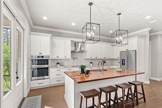 kitchen with wall chimney exhaust hood, sink, wooden counters, stainless steel appliances, and white cabinets