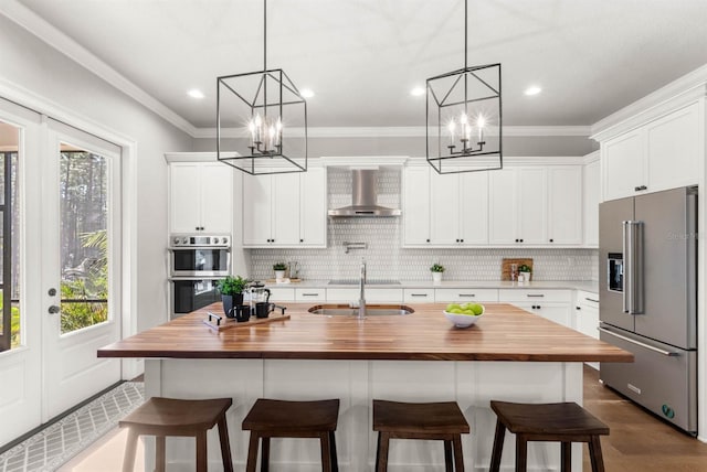 kitchen featuring appliances with stainless steel finishes, white cabinetry, butcher block counters, a kitchen island with sink, and wall chimney exhaust hood