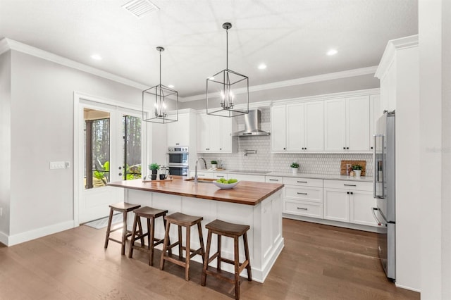 kitchen with wood counters, wall chimney exhaust hood, a kitchen bar, white cabinetry, and an island with sink