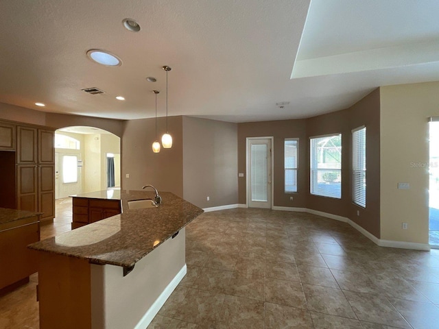 kitchen with a sink, dark stone counters, brown cabinetry, a center island with sink, and pendant lighting