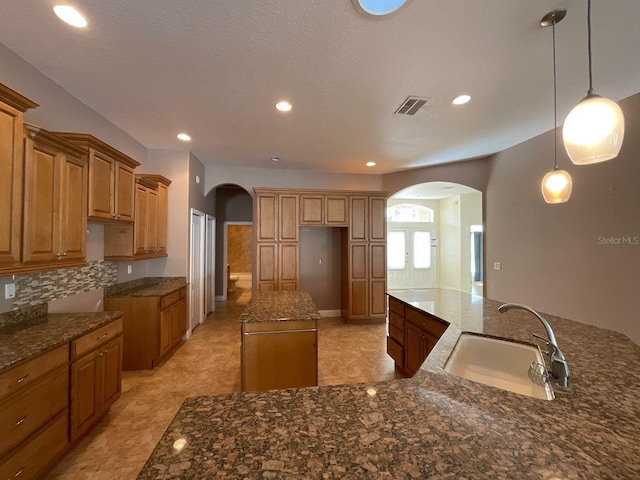 kitchen with sink, hanging light fixtures, a kitchen island, decorative backsplash, and dark stone counters