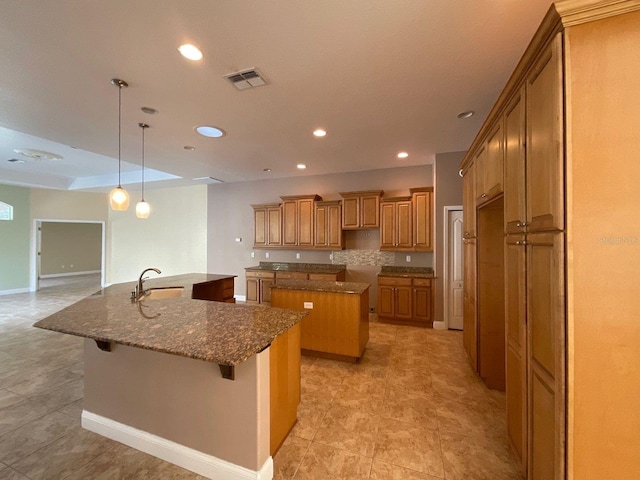 kitchen featuring a kitchen island with sink, a sink, visible vents, brown cabinets, and dark stone counters