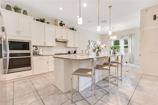 kitchen featuring built in microwave, white cabinets, hanging light fixtures, a kitchen island with sink, and stainless steel oven