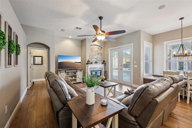 living room with ceiling fan with notable chandelier, light hardwood / wood-style floors, a textured ceiling, a stone fireplace, and french doors