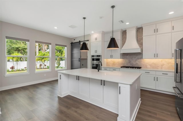 kitchen with custom exhaust hood, decorative light fixtures, backsplash, an island with sink, and white cabinets