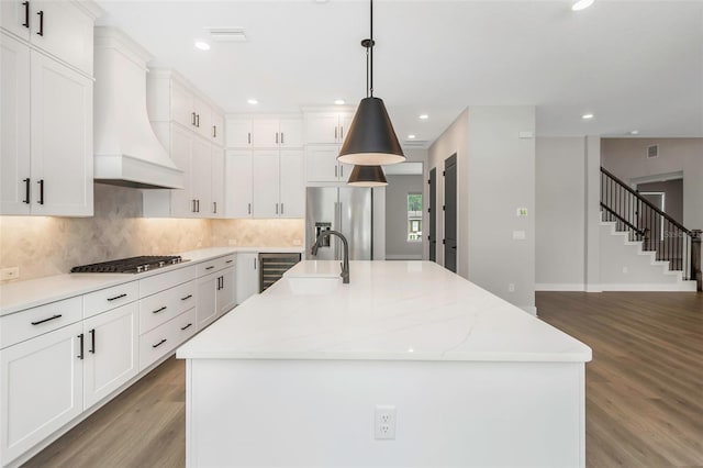 kitchen with white cabinetry, appliances with stainless steel finishes, custom range hood, and a kitchen island with sink