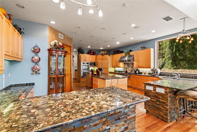 kitchen featuring wall chimney range hood, a kitchen island with sink, hanging light fixtures, stainless steel appliances, and light hardwood / wood-style floors