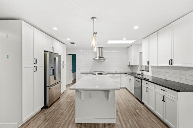 kitchen featuring white cabinets, hanging light fixtures, a center island, stainless steel appliances, and wall chimney range hood