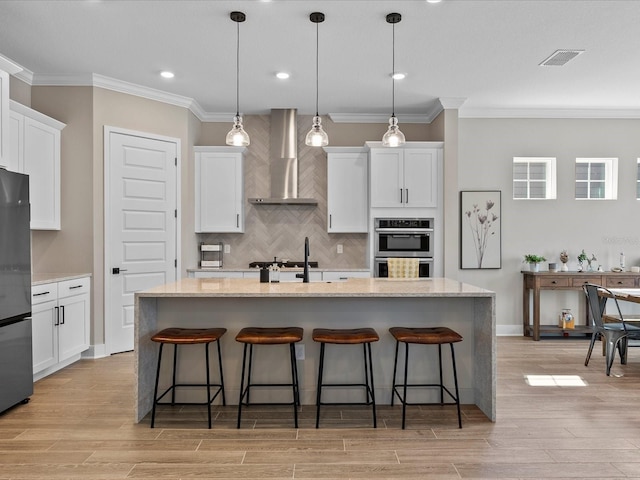 kitchen with an island with sink, decorative light fixtures, white cabinets, and wall chimney exhaust hood