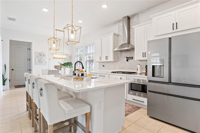 kitchen with pendant lighting, an island with sink, white cabinets, stainless steel appliances, and wall chimney range hood