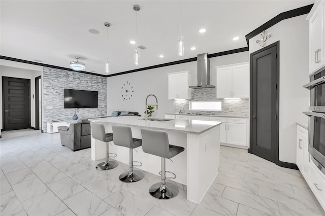 kitchen featuring pendant lighting, white cabinetry, sink, wall chimney range hood, and a center island with sink