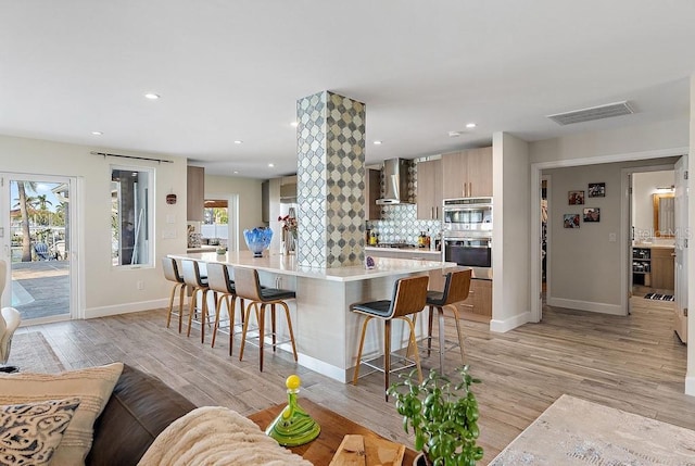 kitchen featuring wall chimney exhaust hood, a kitchen bar, stainless steel appliances, light hardwood / wood-style floors, and backsplash