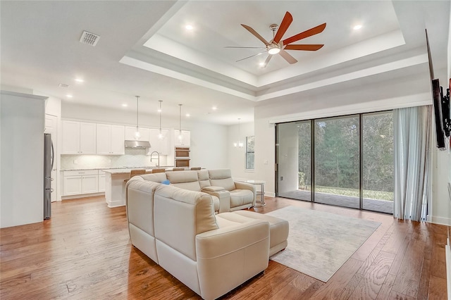 living room with sink, a tray ceiling, light hardwood / wood-style flooring, and ceiling fan