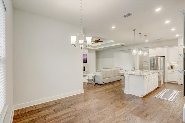 kitchen with stainless steel fridge with ice dispenser, light hardwood / wood-style flooring, hanging light fixtures, an island with sink, and light stone countertops