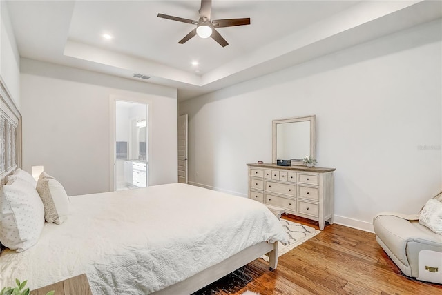 bedroom featuring ceiling fan, ensuite bathroom, a raised ceiling, and light hardwood / wood-style floors