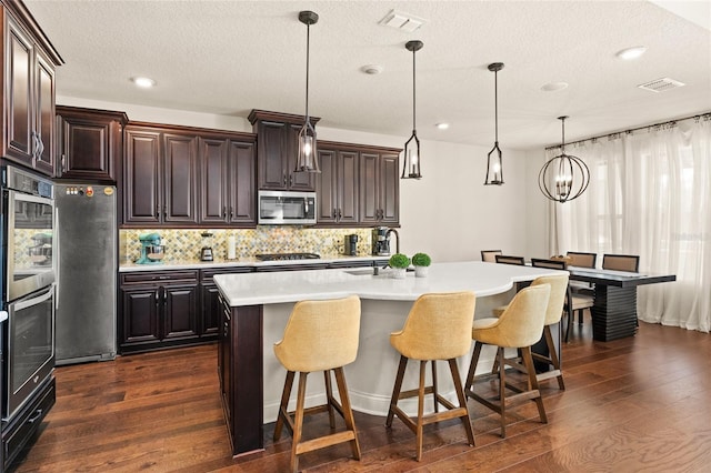 kitchen with dark brown cabinets, hanging light fixtures, appliances with stainless steel finishes, dark hardwood / wood-style floors, and an island with sink