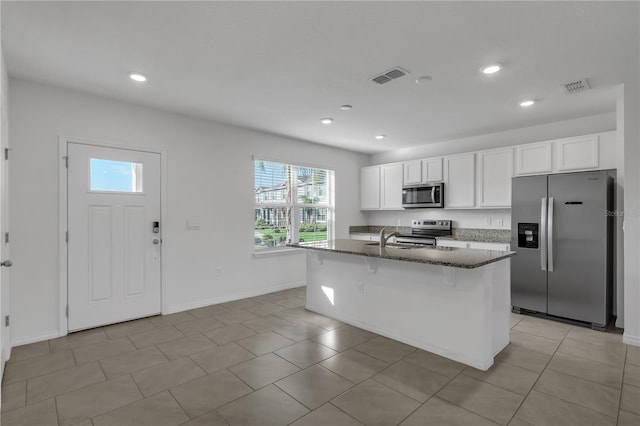 kitchen with dark stone countertops, stainless steel appliances, an island with sink, white cabinets, and a kitchen bar