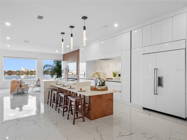 kitchen featuring an island with sink, white cabinets, hanging light fixtures, paneled built in fridge, and a water view