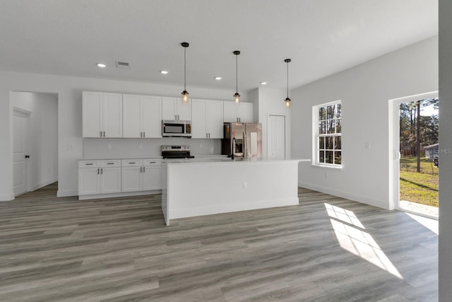 kitchen with appliances with stainless steel finishes, wood-type flooring, white cabinets, a center island with sink, and decorative light fixtures