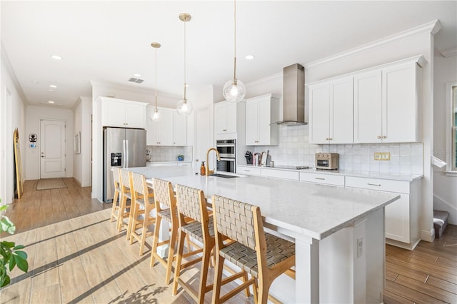 kitchen featuring white cabinets, stainless steel appliances, wall chimney exhaust hood, and a center island with sink