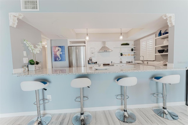 kitchen with pendant lighting, stainless steel fridge, a kitchen bar, kitchen peninsula, and wall chimney range hood