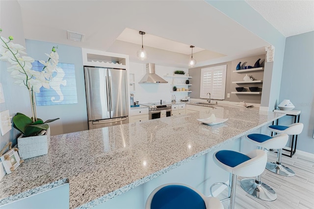 kitchen with appliances with stainless steel finishes, hanging light fixtures, kitchen peninsula, light stone countertops, and wall chimney exhaust hood