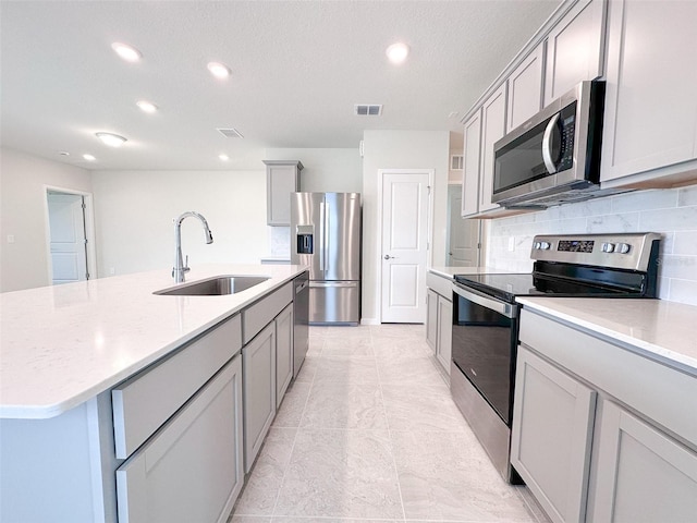 kitchen with sink, appliances with stainless steel finishes, gray cabinetry, backsplash, and an island with sink