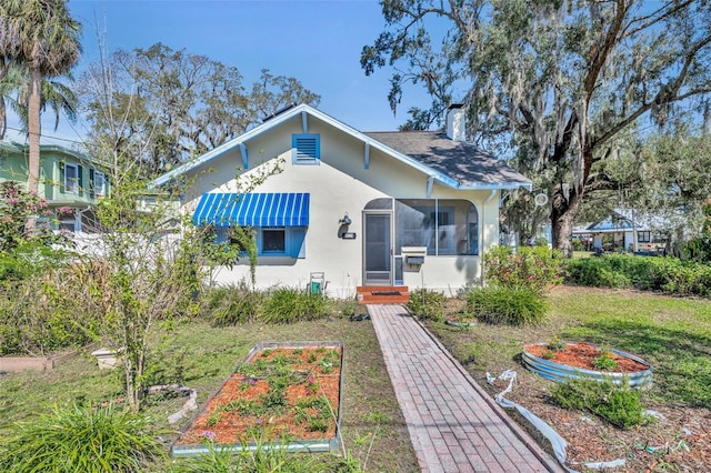 bungalow-style home featuring a vegetable garden, a chimney, stucco siding, a front yard, and a sunroom