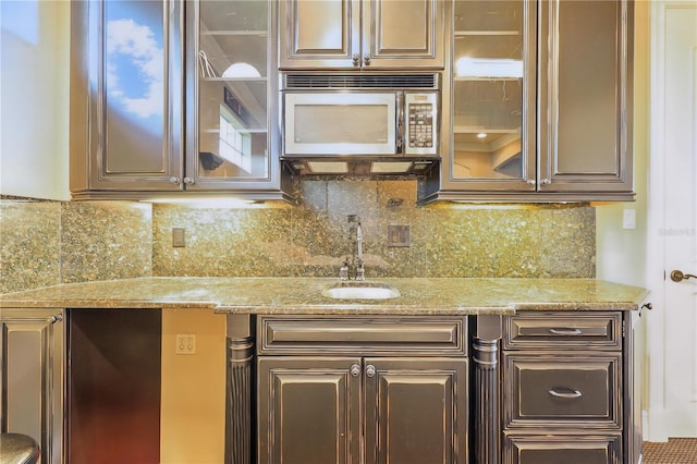 kitchen featuring light stone counters, stainless steel microwave, backsplash, glass insert cabinets, and a sink