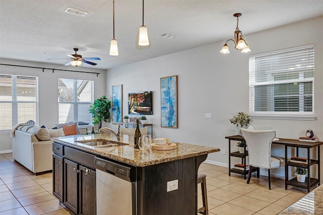 kitchen with light tile patterned floors, dishwasher, hanging light fixtures, light stone counters, and an island with sink