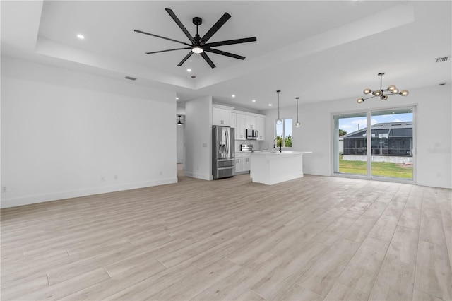 unfurnished living room featuring sink, a tray ceiling, ceiling fan with notable chandelier, and light wood-type flooring