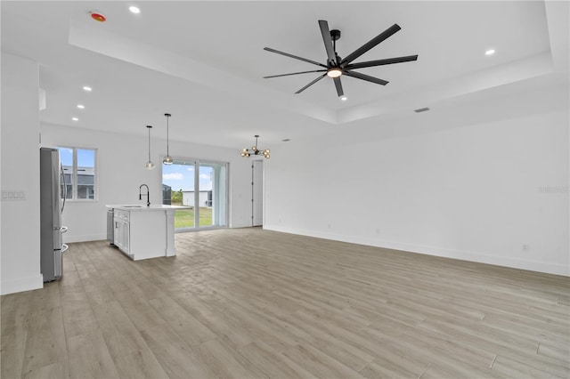 unfurnished living room with sink, ceiling fan with notable chandelier, light wood-type flooring, and a tray ceiling