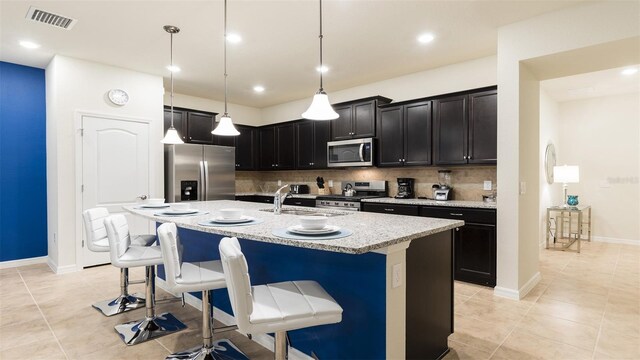 kitchen featuring hanging light fixtures, a center island with sink, stainless steel appliances, light stone countertops, and a kitchen bar