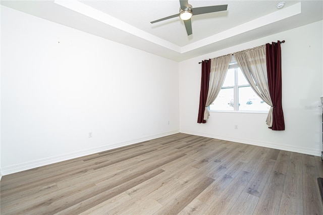 empty room featuring a tray ceiling, ceiling fan, light wood-style flooring, and baseboards