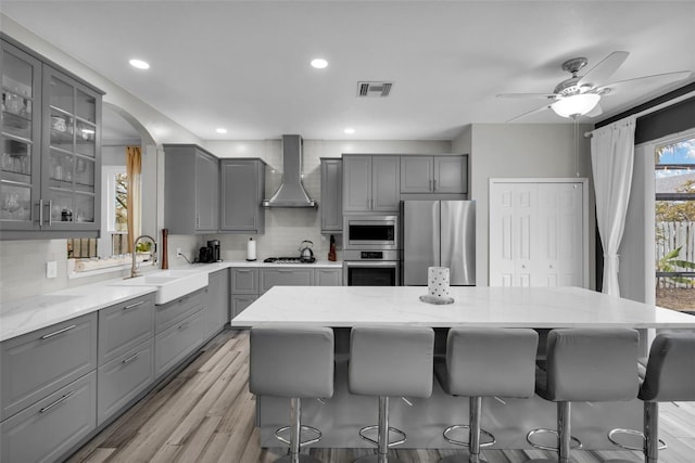 kitchen with sink, appliances with stainless steel finishes, gray cabinetry, a kitchen island, and wall chimney exhaust hood