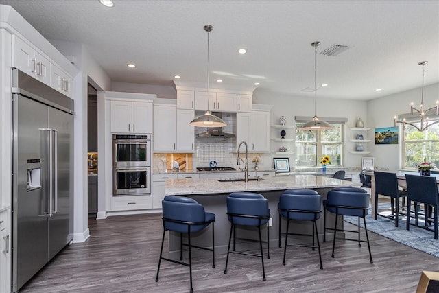 kitchen featuring pendant lighting, white cabinetry, stainless steel appliances, light stone countertops, and an island with sink