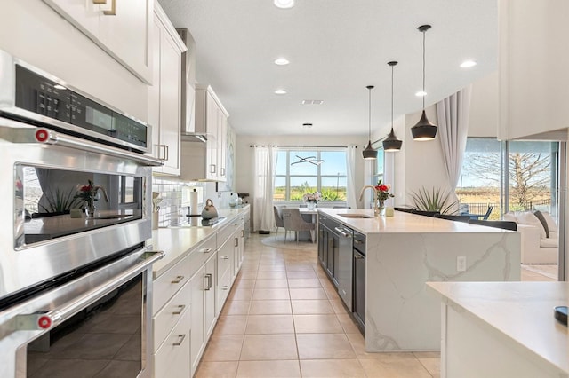 kitchen with double oven, white cabinetry, hanging light fixtures, backsplash, and an island with sink