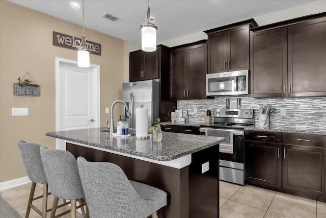 kitchen with stainless steel appliances, a kitchen island with sink, decorative light fixtures, and dark stone counters